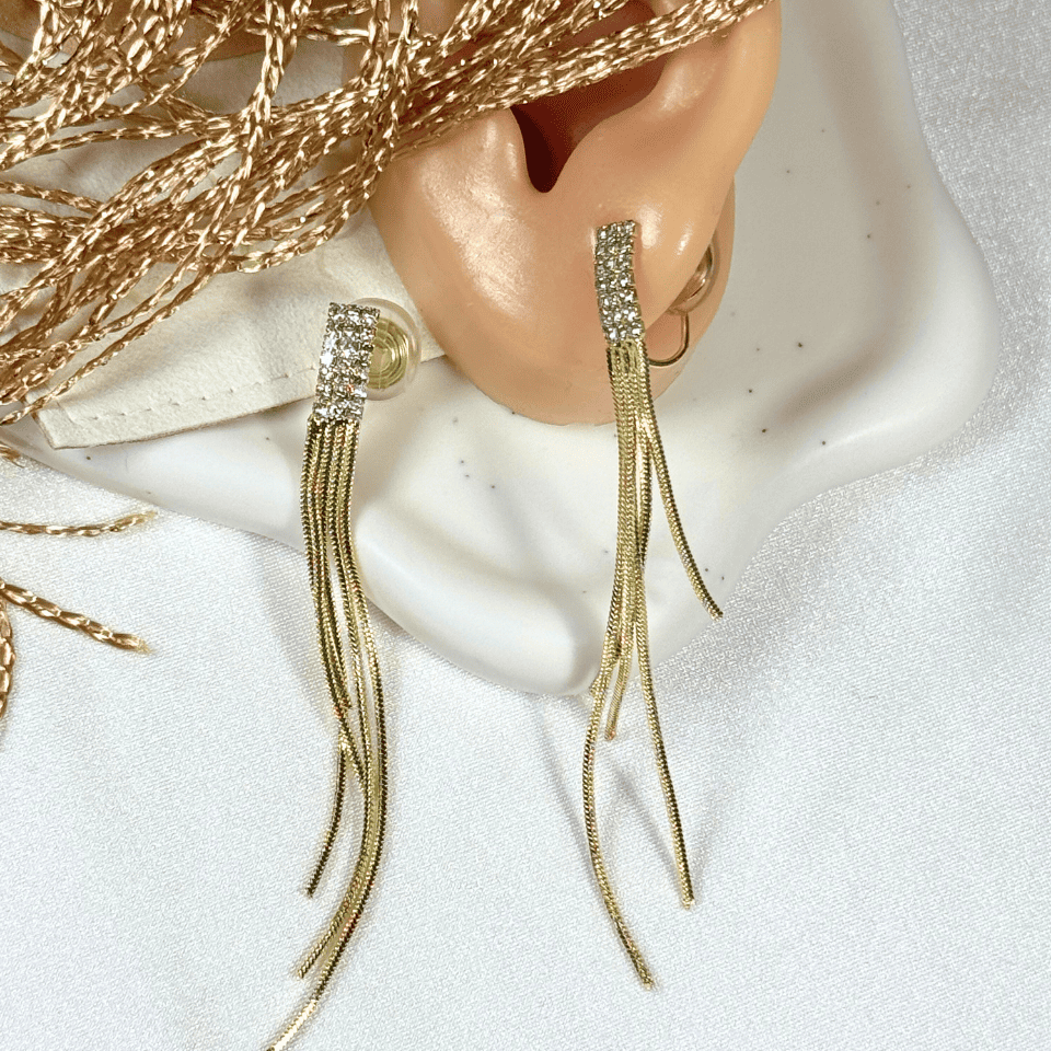 Gold earrings with tassels worn on an ear, with a textured gold background.